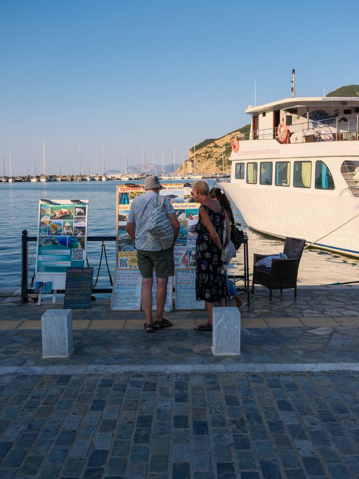 Two adults view excursion options on a sunny day by the port in Skopelos, Greece.